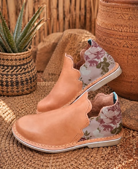 Pair of tan leather childrens boots with floral fabric accents on a woven rug, surrounded by potted succulents and boho decor