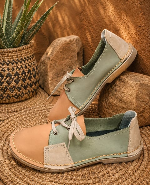 Pair of tan and green leather ankle boots with lace-up front displayed on decorative stones next to a potted succulent plant